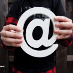 En person, der holder et stort fysisk snabel-a (@) symbol, hvilket illustrerer værdien og prisen ved at have sit eget domæne til en professionel mail til virksomhed.